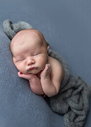 newborn boy posed on steel blue fabric with hands on cheeks.