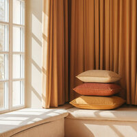 Stack of neutral-toned pillows on a window seat with golden curtains and sunlight streaming through a large-paned window.