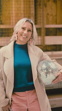 Kirsty in a woolshed with a disco ball under her arm. She is looking relaxed and happy as she smiles at the camera