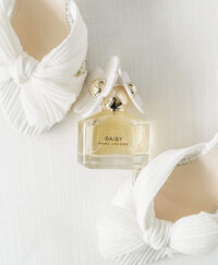 A wedding perfume bottle styled between two white wedding high heels on a white background curated for wedding detail photos