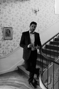 Groom in tuxedo leans on spiral staircase railing with floral wallpaper and antique artwork in the background.