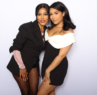 Two women dressed in stylish black-and-white outfits posing together at a photo booth with a white backdrop, smiling and leaning into each other.