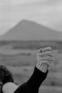 Black and white close-up of a couple holding hands, engagement ring in focus, with a blurred volcanic mountain in the background — captured in Hella, Iceland by Gaby, Destination Elopement Photographer.