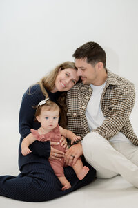 A pregnant mother sits on the ground with her legs over her husband as she's laughing. Her husband is holding her legs, looking at her smiling.