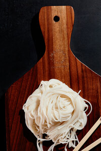 A minimalist and clean image of fresh noodles artfully arranged on a wooden cutting board. Photographed by Mica McCook, this composition highlights the simplicity and beauty of traditional ingredients, emphasizing texture and form.