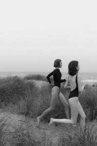 Two women in sleek swimwear running through sandy dunes near the ocean, showcasing a minimalist and elegant beach scene.