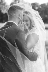 Bride and groom stand under veil while groom kisses bride