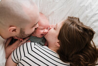 Infant smiling on white bed cover while parents kiss her cheeks simple infant photography at home