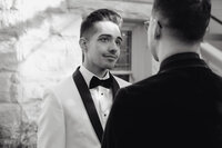 A groom in a white tuxedo gazes at his partner during their intimate wedding ceremony, captured in a cinematic and documentary wedding photography style.