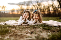 Two girls lay on blanket facing camera in field with trees and sunset behind them with face on hands while smiling at camera.