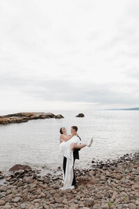 A person holding up their partner as they stand on a rocky shoreline 