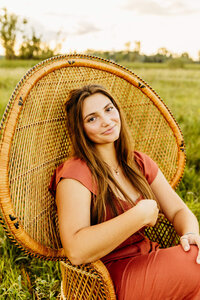 beauitiful senior girl sitting on a peacock chair while playing with her hair captured by Ashley Kalbus