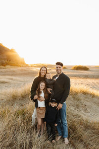 A set of parents and their children smile at the camera while photographed by family photographer lucy jane in christchurch at their favourite beach