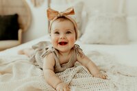 A baby with a headband and bow lies on a cozy bed, smiling and wearing a beige outfit.