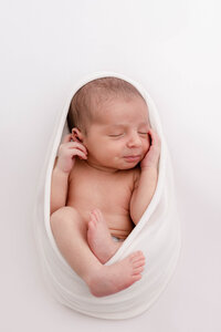 Newborn Baby wrapped in a white swaddles with hands, chest, and legs showing. Sleeping peacefully on a white backdrop.