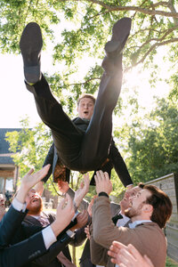 A groom is joyfully tossed into the air by a group of groomsmen during a wedding celebration. His expression is one of surprise and excitement as he soars above them.