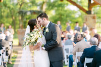 A Dallas-Fort-Worth-Wedding-Photographer-and-videographer captures a newlywed couple sharing a kiss at their outdoor ceremony, surrounded by guests seated under a wooden structure on a sunny day.