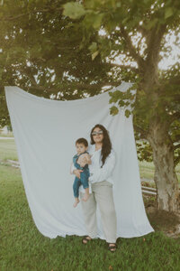 Family Photoshoot: Mother Poses with Son in Her Arms Outdoors, Against a White Blanket Background.