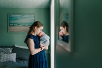 mom holding newborn son in front of a window with their reflection being cast in the frame of the foreground