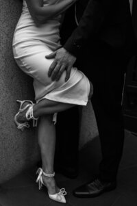 Black and white artistic photo of a couple embracing, focusing on their legs and shoes, with the woman wearing a flowing white dress and heels, captured in an elegant engagement session by a professional photographer in Portland.
