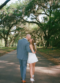 Couple in suit and white dress lean in for a kiss under arch of old trees