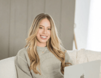 Young woman with long blonde hair, wearing a light gray sweatshirt, relaxing on a cozy couch with a MacBook in front of her, smiling at the camera in a softly lit room.