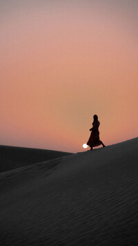 Woman walking across Sahara dunes at sunset with long shadow