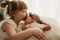 a toddles sister who’s kissing and hugging a newborn baby that is sleeping
