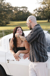 Couple sharing a tender moment beside a vintage white car in a sunlit field, captured in a documentary-style portrait session.