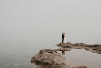 bride and groom standing on rock next to water