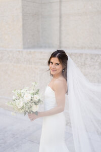 A bride in a white wedding dress smiles softly through her veil on the grounds of the Ohio Statehouse