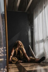 Andrea Cakmar sits on the floor of a sunlit room, leaning back against a wall with a relaxed expression. She wears a tan outfit, and the room has large windows with sheer curtains allowing natural light to flood in.