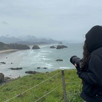 Danielle Rose Photography holding her camera and looking out at the breathtaking view from Ecola State Park in Oregon, capturing the essence of travel wedding photography amidst stunning coastal scenery.