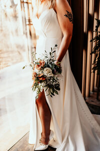 Details of the brides dress, flowers and shoes as she looks out the large glass pane of the glass chapel