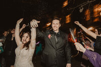 A newlywed couple with their hands up as they walk in between their wedding guests 