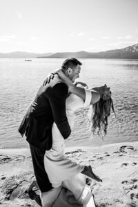 Black and white photo of a joyful couple eloping on a sandy beach at Lake Tahoe, with the groom dipping the bride in a playful embrace by the water.