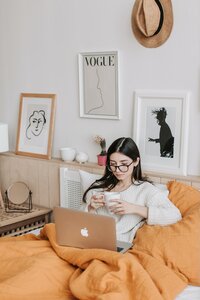 Woman sitting in bed working from home with a laptop on her lap and tea mug in her hands