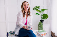 White woman sitting in front of a white background near a table with a green leaf plant next to her. She is in a white shirt and holding a black and white coffee cup.