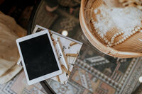 A tablet with a blank screen rests on a notebook and pen atop a glass table, next to a wicker basket filled with pearl necklaces and decor. Perfect for outlining your next white label design course, with a patterned rug beneath the table.