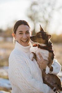 A smiling white woman with dark hair tied back, wearing a white turtleneck sweater and earrings. She is holding a small brown and white dog with perky ears and a red bandana. They are outdoors, with a softly blurred natural background indicating it may be late afternoon due to the warm lighting. The dog is looking affectionately at the woman, suggesting a close bond between them.