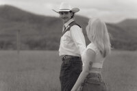 A black and white photo of a young man in a cowboy hat leading his girlfriend through a field with mountains in the background in Northeast TN