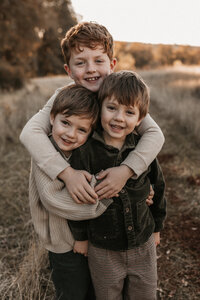 A big brother with red hair wrapping his arms around his two little brothers in an El Dorado Hills, CA field. 