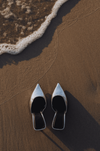 A pair of silver high-heeled shoes sit on a sandy beach with a gentle wave lapping at the shore. The image evokes a sense of luxury and relaxation.