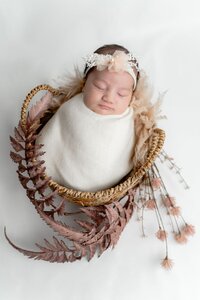 Newborn baby swaddled in a soft white wrap, wearing a delicate peach floral headband, sleeping in a wicker basket decorated with dried ferns.