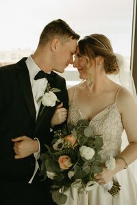 A bride and groom in formal attire share an intimate moment during their full service wedding planning, with the bride holding a bouquet of white and green flowers.