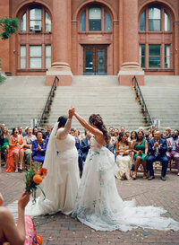 Film photo of two brides raising their hands in celebration during an outdoor wedding ceremony at the Multicultural Arts Center in Cambridge, MA — captured by a film wedding photographer documenting inclusive love stories.