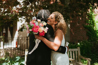 Bride hugging groom during first look, smiling while holding a bright bouquet with coral, yellow, and blush-toned blooms.