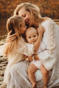 A mom hugging her two little girls while the youngest looks at the camera at the lake in Canandaigua, NY