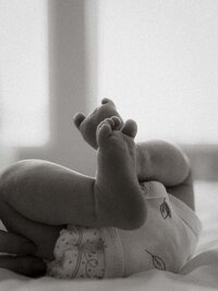 Black and white photo of newborn baby’s feet lifted gently in the air during a quiet in-home session, capturing the tenderness of early days.