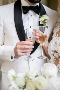 A couple in formal wedding attire, with the groom in a white suit and black bow tie, and the bride in a lace dress, holding champagne glasses for a toast. A floral boutonniere is visible on the grooms lapel.